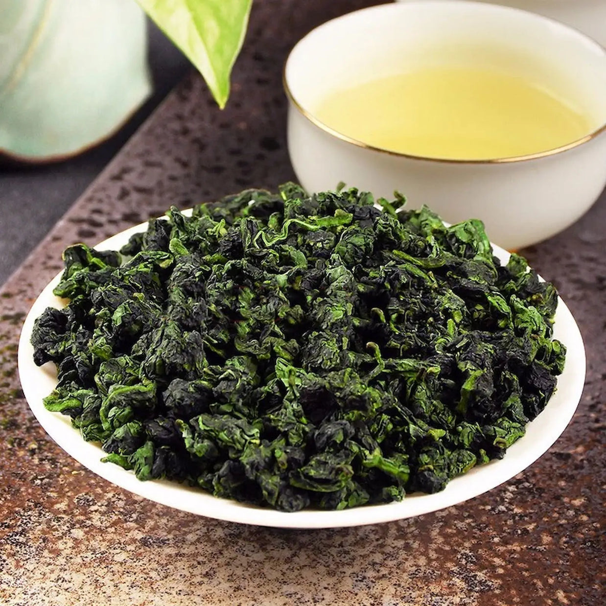 Freshly rolled Anxi Tie Guan Yin oolong tea leaves in a white dish, with a cup of light golden brewed tea in the background. Premium Chinese oolong known for its floral aroma and smooth, refreshing taste, ideal for Gongfu tea ceremonies.
