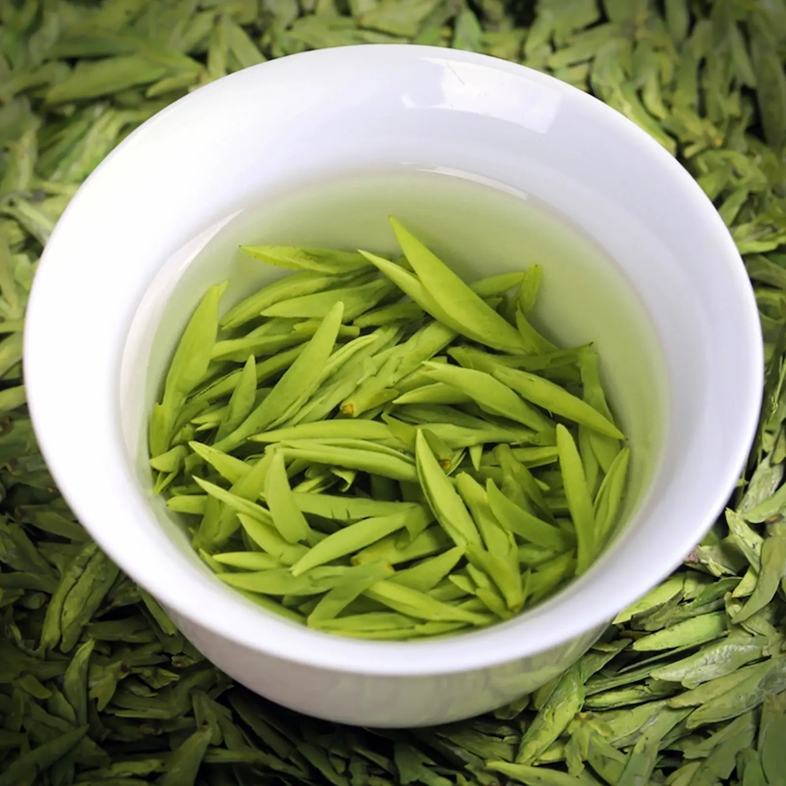 Freshly steeped Ming Qian Longjing green tea leaves in a white porcelain cup, showcasing vibrant green buds and clear tea liquor. Premium pre-Qingming Dragon Well tea from Hangzhou, prized for its delicate aroma and mellow taste.
