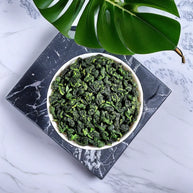 Premium Anxi Tie Guan Yin oolong tea leaves in a white bowl, displayed on a marble background with a green leaf accent. High-quality Chinese oolong known for its vibrant color, floral aroma, and fresh taste, perfect for tea connoisseurs.
