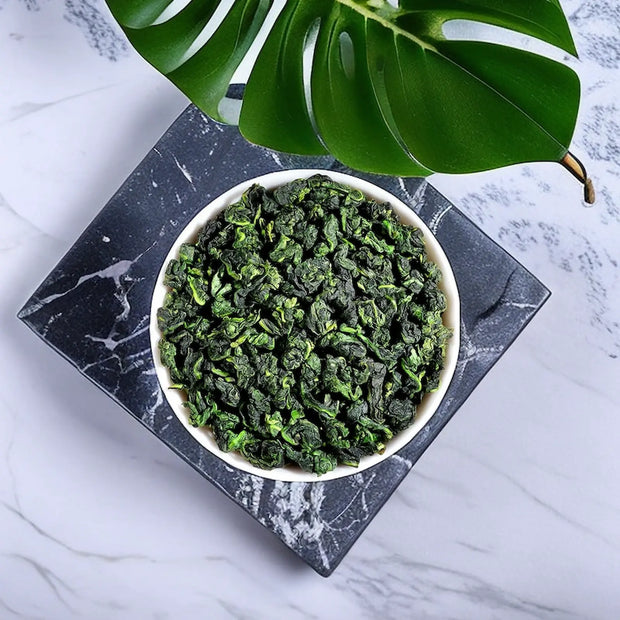 Premium Anxi Tie Guan Yin oolong tea leaves in a white bowl, displayed on a marble background with a green leaf accent. High-quality Chinese oolong known for its vibrant color, floral aroma, and fresh taste, perfect for tea connoisseurs.
