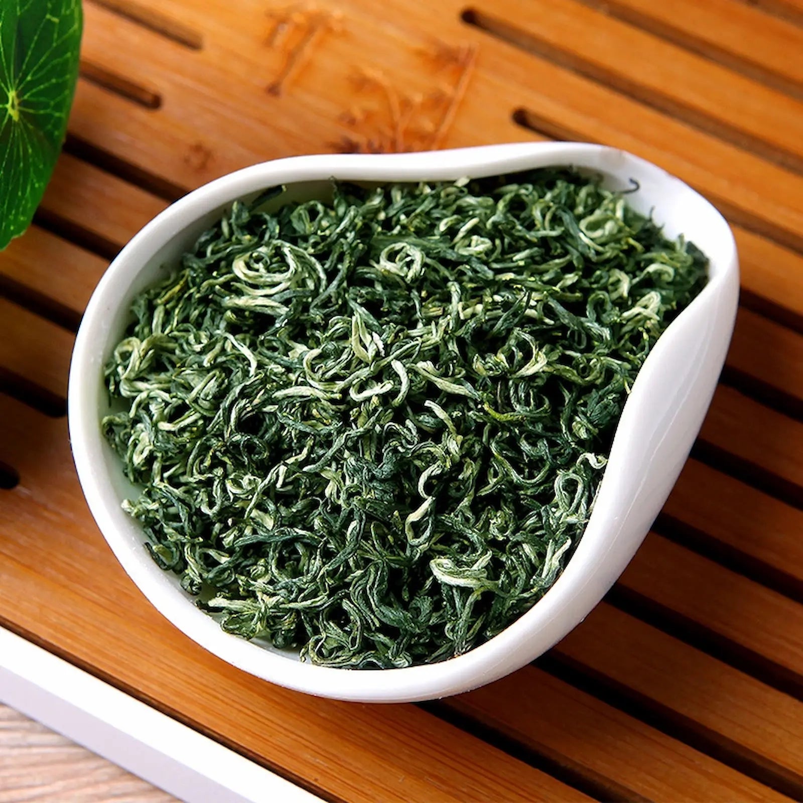 Premium Bi Luo Chun green tea leaves in a white porcelain dish, featuring delicate spiral shapes and fine white hairs. Authentic Chinese spring tea from Dongting Mountain, prized for its fresh aroma and mellow taste.
