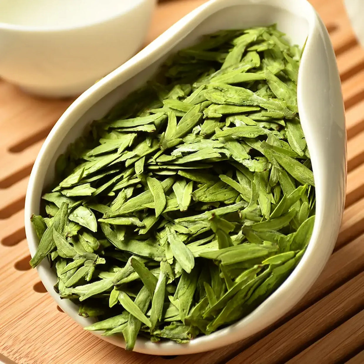 Premium West Lake Longjing green tea leaves in a white porcelain dish, featuring flat, vibrant green buds. Authentic Xi Hu Dragon Well tea from Hangzhou, prized for its fresh aroma and delicate, mellow flavor.
