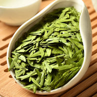 Premium West Lake Longjing green tea leaves in a white porcelain dish, featuring flat, vibrant green buds. Authentic Xi Hu Dragon Well tea from Hangzhou, prized for its fresh aroma and delicate, mellow flavor.
